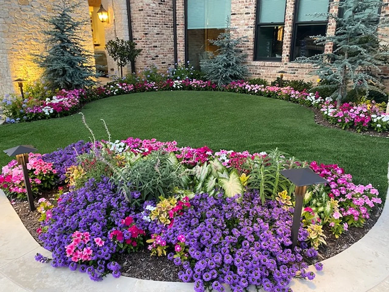 Curved bed of purple petunias and pink blooms beside a brick home