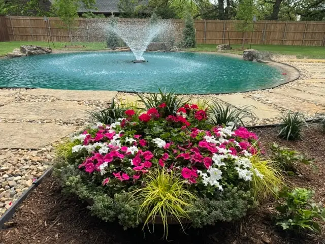 Round bed of pink and white vinca in front of a pond with a fountain