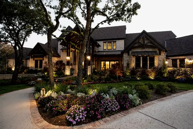 Stone home with lit planting beds and uplit oaks along a curved drive at dusk